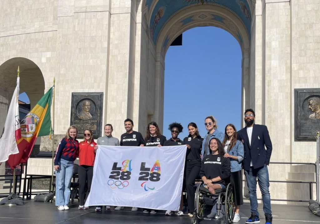 A group of athletes and organisers stand on a stage in front of the Los Angeles Memorial Coliseum arches, holding an “LA 28” banner for the 2028 Olympic and Paralympic Games venue announcement.
