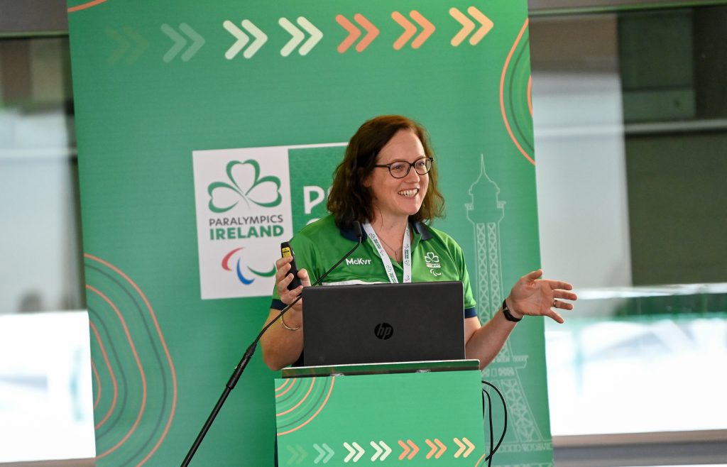 Neasa Russell stands at a podium in front of a green Paralympics Ireland backdrop, smiling as she speaks and gestures while holding a clicker at the Paris 2024 brand launch.