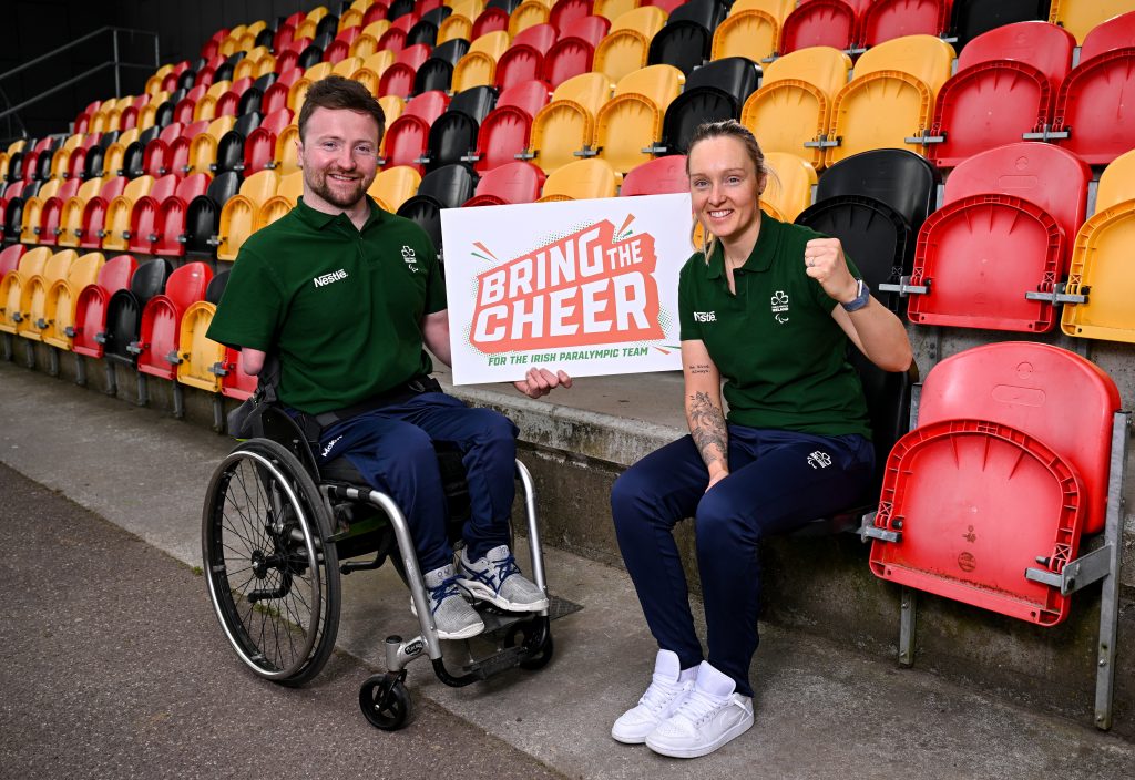 Colin Judge (left) and Richael Timothy (right) sit in stadium seats holding a “Bring the Cheer” sign, with Colin in a wheelchair and Richael raising a fist.