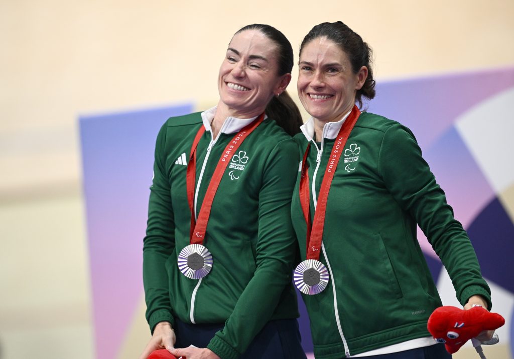 Eve McCrystal and Katie George Dunlevy stand closely together smiling and wearing their green kit with a medal around each of their necks