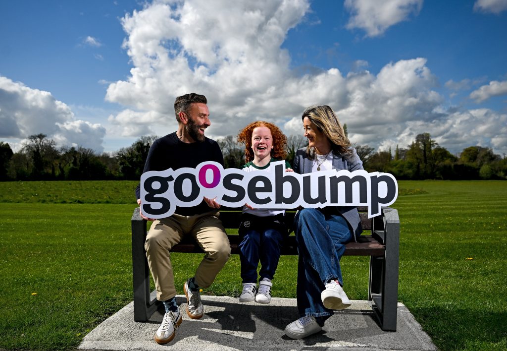 Stephen McNamara (left) Dearbhaile Brady (centre) and Sarah Love (right) sitting on a bench on a sunny day in front of a green field holding a sign saying Goosebump. The group are laughing.
