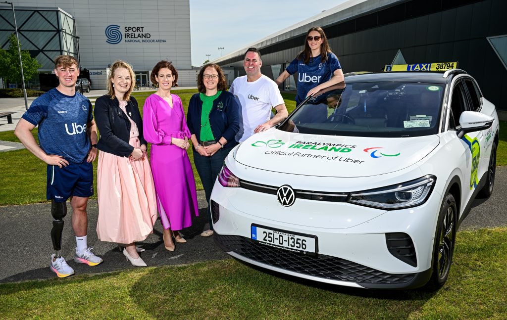 Six people pose beside a white Uber-branded Paralympics Ireland taxi outside the Sport Ireland National Indoor Arena. From left: Paralympian Tiarnán O’Donnell, (wearing a navy Uber t-shirt) Lisa Clancy, Hildegarde Naughton, Neasa Russell, Kieran Harte, and Róisín Ní Riain (wearing a navy Uber t-shirt).
