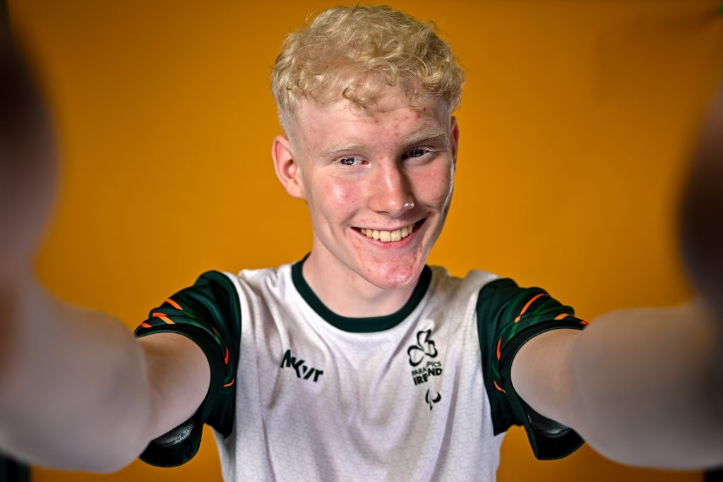 Damien Moyles, Para Athletics, poses for portrait during the European Youth Games 2025 Team Photo Shoot at the Sport Ireland Institute on the Sport Ireland Campus, Dublin. Smiling with both hands up to camera. Close up shot. Orange background white t shirt