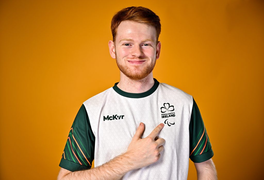 Sean Geoghegan, a para table tennis athlete, poses for a portrait at the European Youth Games 2025 team photo shoot, pointing to the Paralympics Ireland logo on his kit.
