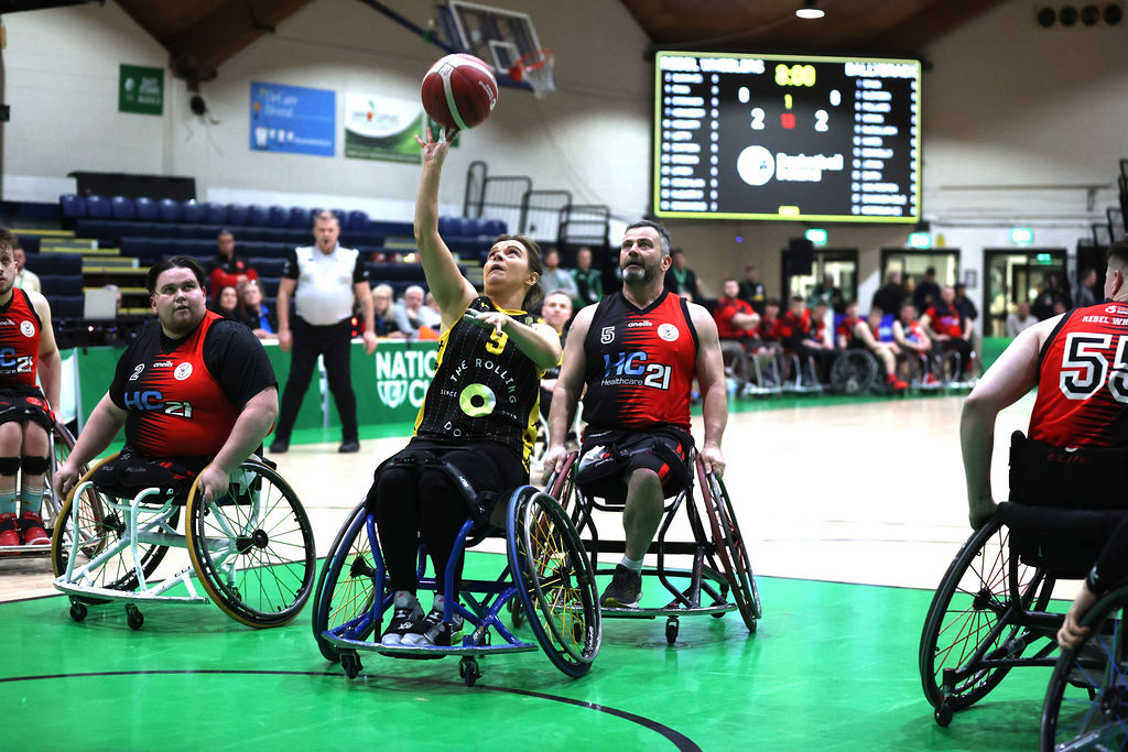 A female player wearing black with yellow trim is surrounded by opposition players wearing red and black as she catches the ball