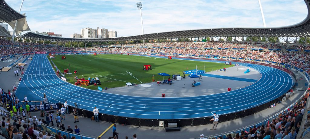 Wide panoramic view of Stade Charléty in Paris, showing a blue running track surrounding a grass infield, with packed spectator stands and athletics equipment set up around the stadium.