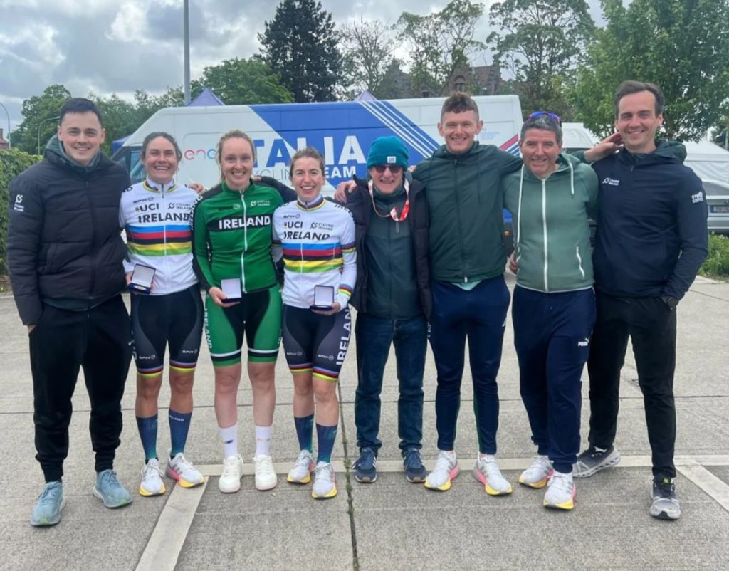 A group photo of the Irish Para Cycling Team and staff with Katie George Dunlevy and Linda Kelly in their white kit, with Richael Timothy in a green kit, each holding their medals