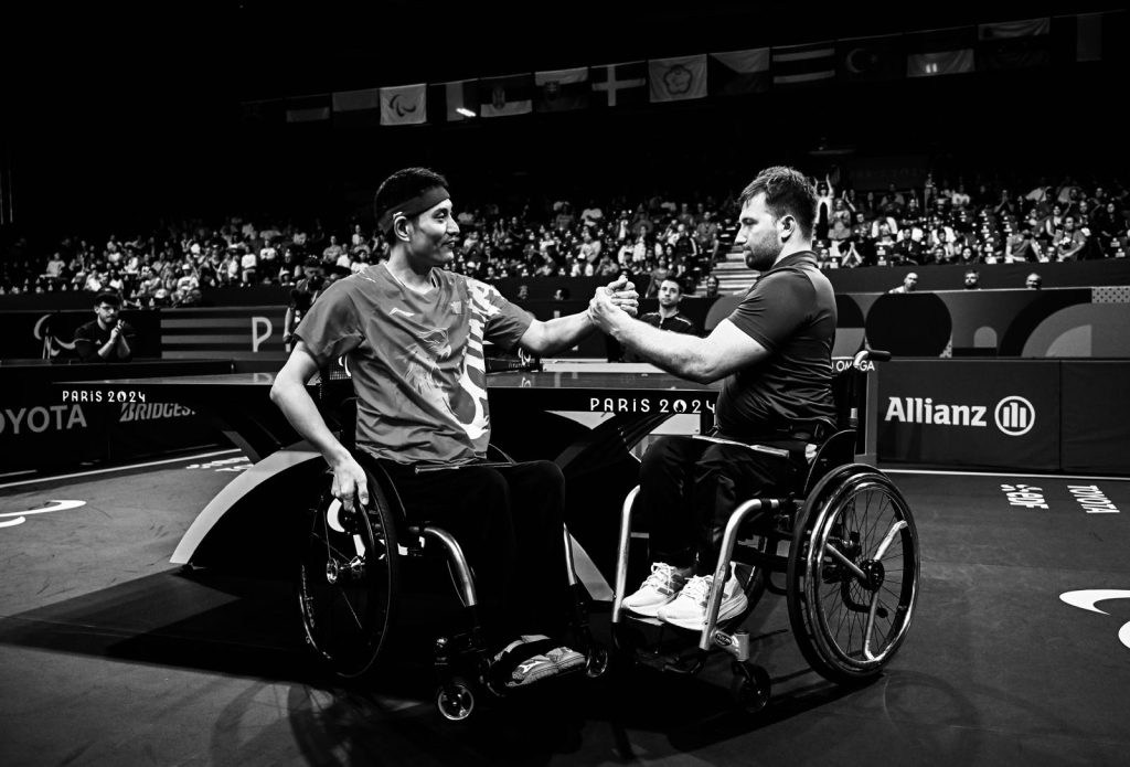 In a black-and-white image, Colin Judge of Ireland (right) shakes hands with Feng Panfeng of China while seated in their wheelchairs after their men’s singles MS3 match at the Paris 2024 Paralympic Games.